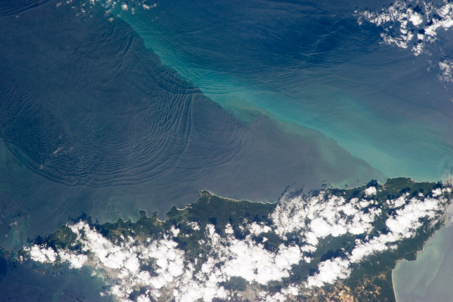 A photograph taken from the International Space Station showing a series of subtle, interacting arcs in the southeastern Caribbean Sea over the northern coast of the island of Trindad. These are known as “internal waves,” the surface manifestation of slow waves that move tens of meters beneath the sea surface. (Courtesy of NASA)