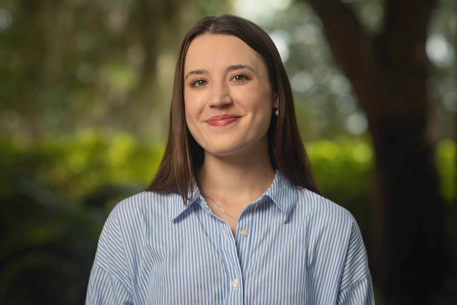 Headshot of Lili Boss in a blue stripped shirt with a blurred background of trees. 