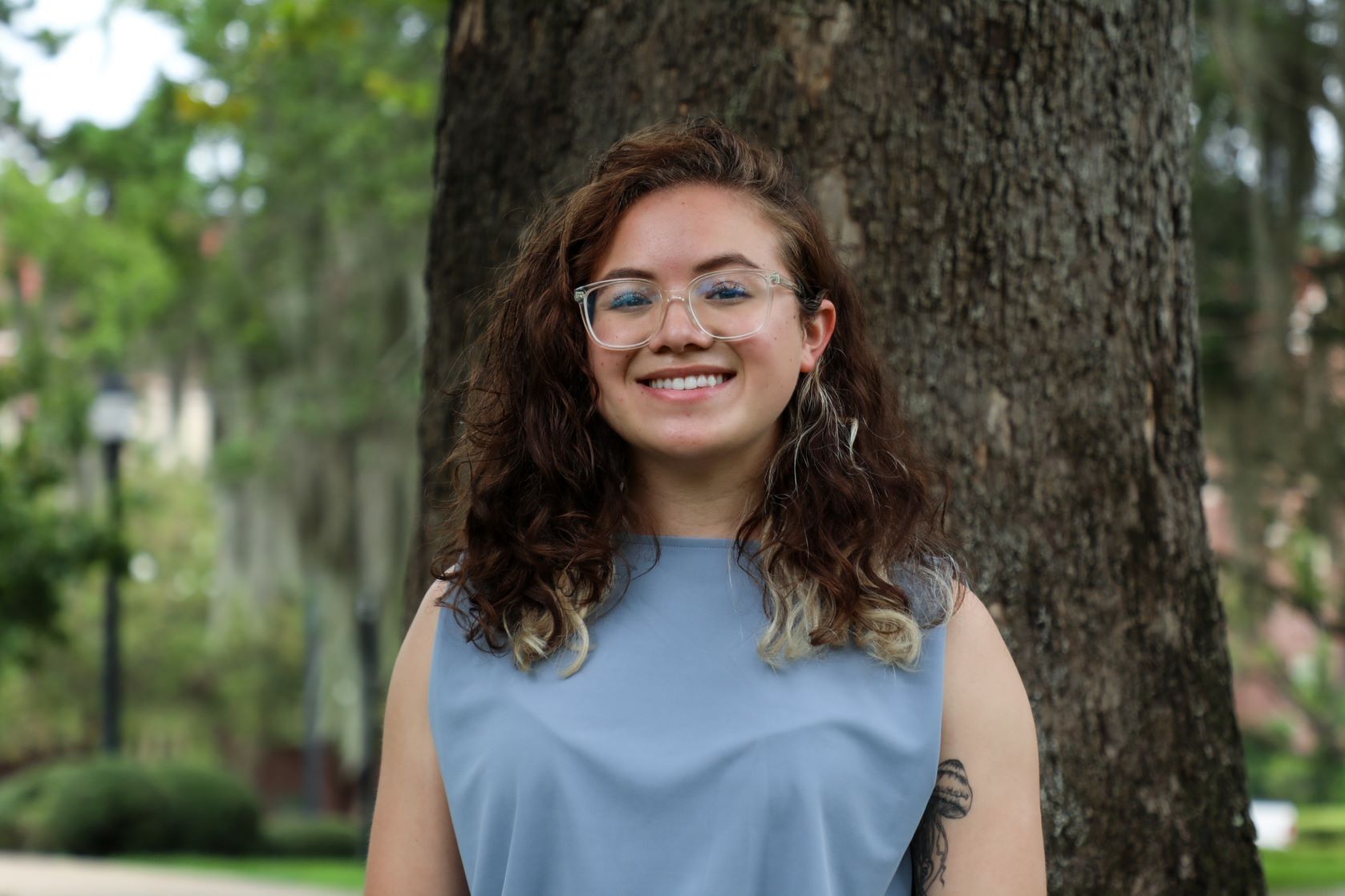 This is a headshot of Andrea in a blue tank top standing in front of a tree. 