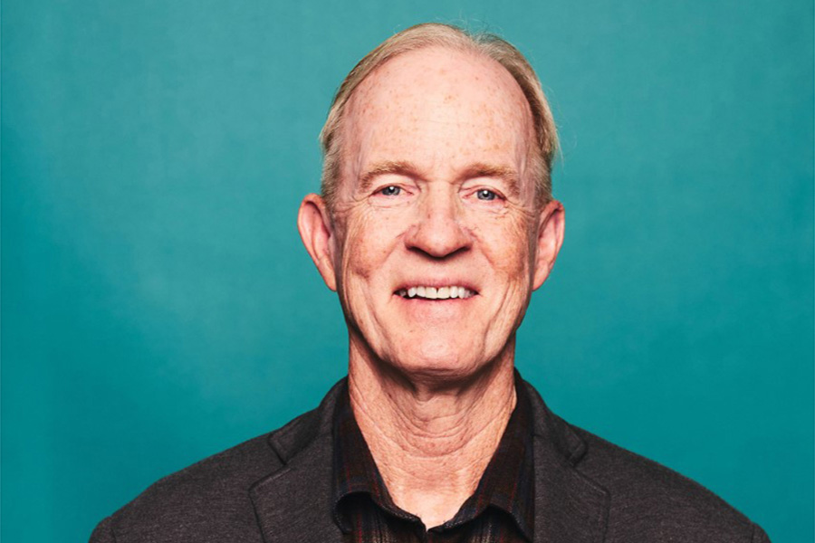 This is a headshot of Mark Powell in a black shirt in front of a blue background. 